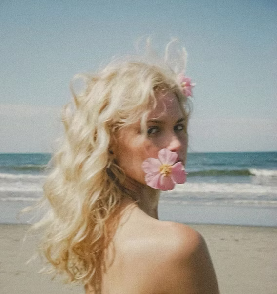 blonde girl at the beach with pink flowers in her hair and mouth